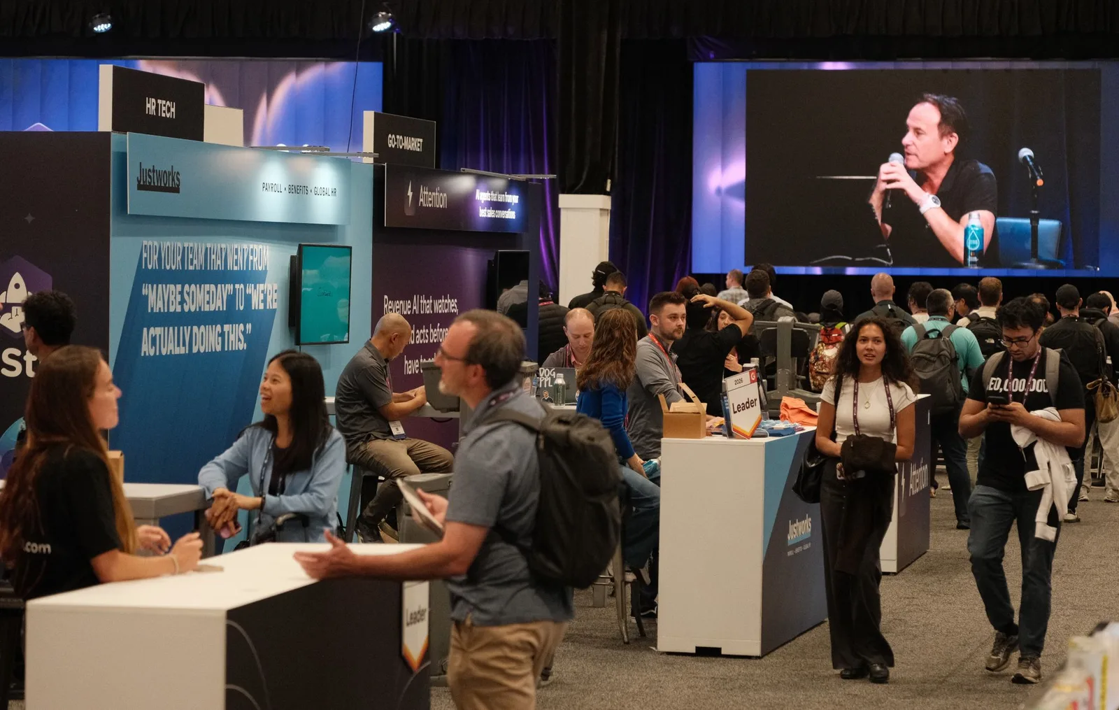 Wide shot of the SaaStr AI show floor with attendees moving between booths and a speaker on the main stage screen